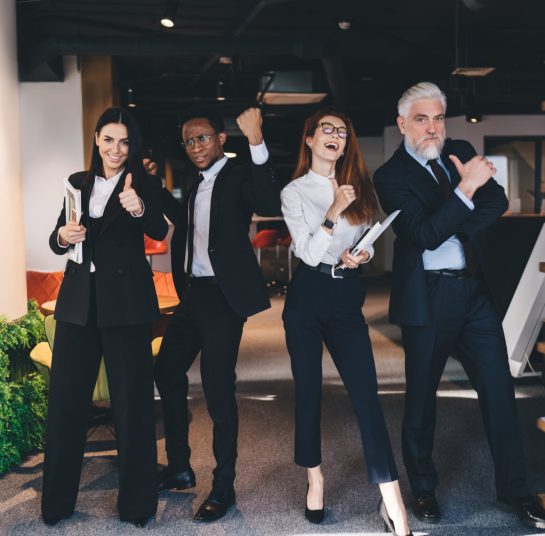 Group of cheerful multiracial coworkers in formal suits standing in office cafeteria with documents near bookshelves and showing thumbs up while celebrating successful deal